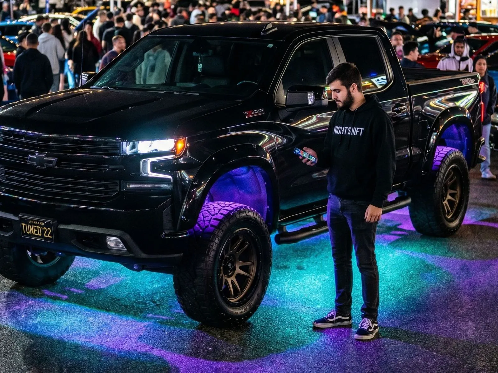 The owner of a 2022 Chevrolet Silverado stands by his vehicle at a car show at nighttime and uses his phone to control his LED underglow system.